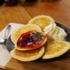 Sur une planche de présentation en bois et marbre, prend place une assiette de pancakes recouverts de gelée de fraises au safran avec un peu de chantilly. En arrière plan, sur une planche ronde en bois, le pot de gelée de fraises au safran