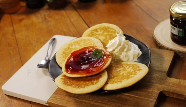 Sur une planche de présentation en bois et marbre, prend place une assiette de pancakes recouverts de gelée de fraises au safran avec un peu de chantilly. En arrière plan, sur une planche ronde en bois, le pot de gelée de fraises au safran