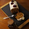 Sur une ardoise entourée de bois, un cake à la carotte recouvert de chantilly et de décor à la gelée de fleurs d'Hibiscus au safran prêt à être dégusté. Au premier plan, à gauche, un pot de safran.