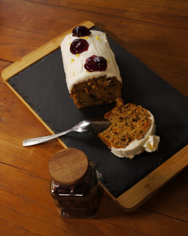 Sur une ardoise entourée de bois, un cake à la carotte recouvert de chantilly et de décor à la gelée de fleurs d'Hibiscus au safran prêt à être dégusté. Au premier plan, à gauche, un pot de safran.