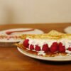 Au premier plan, sur une assiette, une crêpe fourrée de gelée de pétales de rose au safran, avec des framboises et de la chantilly. derrière, sur une planche ronde en bois, quelques feuilles de roses éparpillées et une cuillère en porcelaine rouge, sur la droite le pot de gelée de pétales de rose au safran est présenté sur une planche ronde en bois