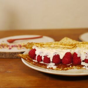 Au premier plan, sur une assiette, une crêpe fourrée de gelée de pétales de rose au safran, avec des framboises et de la chantilly. derrière, sur une planche ronde en bois, quelques feuilles de roses éparpillées et une cuillère en porcelaine rouge, sur la droite le pot de gelée de pétales de rose au safran est présenté sur une planche ronde en bois