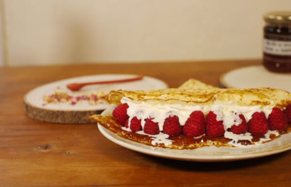 Au premier plan, sur une assiette, une crêpe fourrée de gelée de pétales de rose au safran, avec des framboises et de la chantilly. derrière, sur une planche ronde en bois, quelques feuilles de roses éparpillées et une cuillère en porcelaine rouge, sur la droite le pot de gelée de pétales de rose au safran est présenté sur une planche ronde en bois