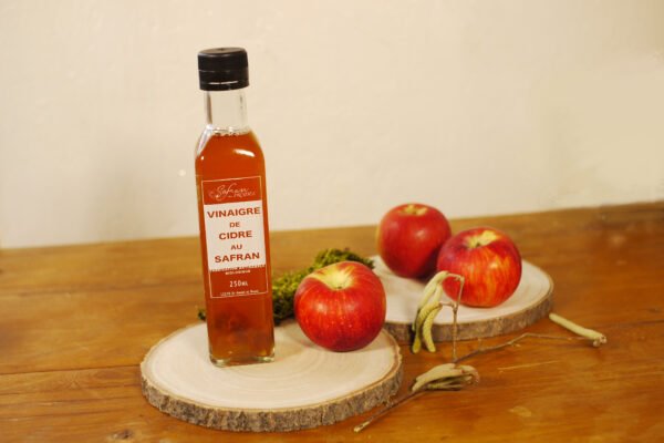 Sur deux planches rondes en bois, prennent place une bouteille de vinaigre de cidre au safran, trois pommes, des chatons de noisetier et un peu de mousse en arrière plan.