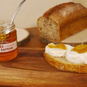 Sur une table en bois, une tartine est recouverte de deux rondelles de chèvre surmontées d'un peu de gelée de acidulée de safran. Derrière la tartine, sur une planche ronde en bois, se trouve un pain en partie coupé. Le pot de gelée acidulée de safran se trouve sur la gauche de la tartine et est ouvert avec une cuillère dedans.