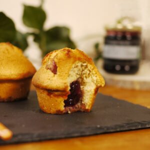 Au premier plan, sur une ardoise sont présentés deux muffins fourrés à la gelée de fleurs de coquelicot au safran, à côté d'une cuillère en porcelaine jaune. En arrière plan, une branche de lierre pour décorer et à droite, sur une planche ronde en bois, le pot de gelée de fleurs de coquelicot au safran.