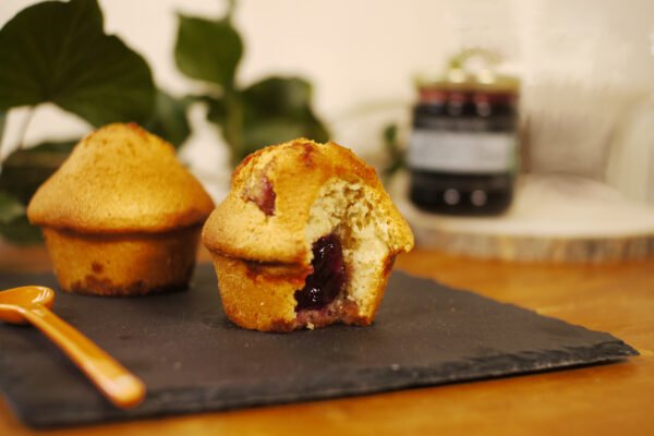 Au premier plan, sur une ardoise sont présentés deux muffins fourrés à la gelée de fleurs de coquelicot au safran, à côté d'une cuillère en porcelaine jaune. En arrière plan, une branche de lierre pour décorer et à droite, sur une planche ronde en bois, le pot de gelée de fleurs de coquelicot au safran.
