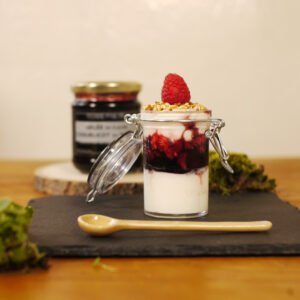 Au premier plan, sur une ardoise est posé une verrine sucrée avec de la gelée de fleurs de coquelicot au safran et des franboises, sur un lit de mascapone. des graines de sésame et une framboise terminent la composition. Devant une cuillère en porcelaine jaune prend place. En arrière plan, sur une planche ronde en bois, le pot de gelée de fleurs de coquelicot au safran.