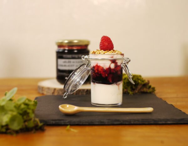 Au premier plan, sur une ardoise est posé une verrine sucrée avec de la gelée de fleurs de coquelicot au safran et des franboises, sur un lit de mascapone. des graines de sésame et une framboise terminent la composition. Devant une cuillère en porcelaine jaune prend place. En arrière plan, sur une planche ronde en bois, le pot de gelée de fleurs de coquelicot au safran.