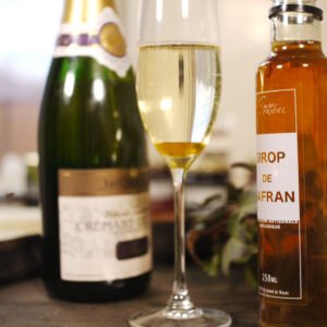 Sur un tonneau de bois décoré de lierre, on trouve au premier plan une flûte à champagne avec dans le fond du sirop de safran et au dessus un pétillant. Sur la droite, prend place la bouteille de sirop de safran et derrière, une bouteille de pétillant.